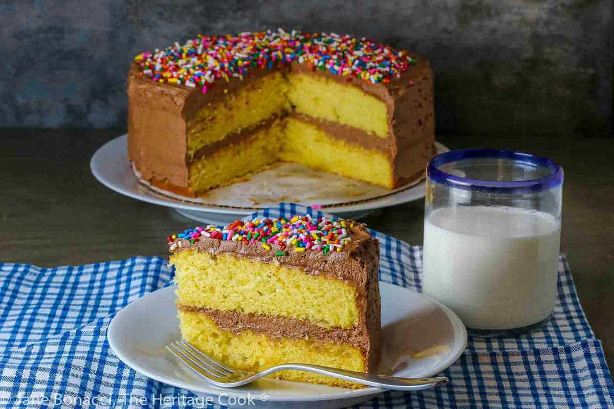 A yellow layer cake with chocolate frosting and colorful sprinkles, served with a slice on a plate beside a glass of milk on a blue checkered tablecloth.