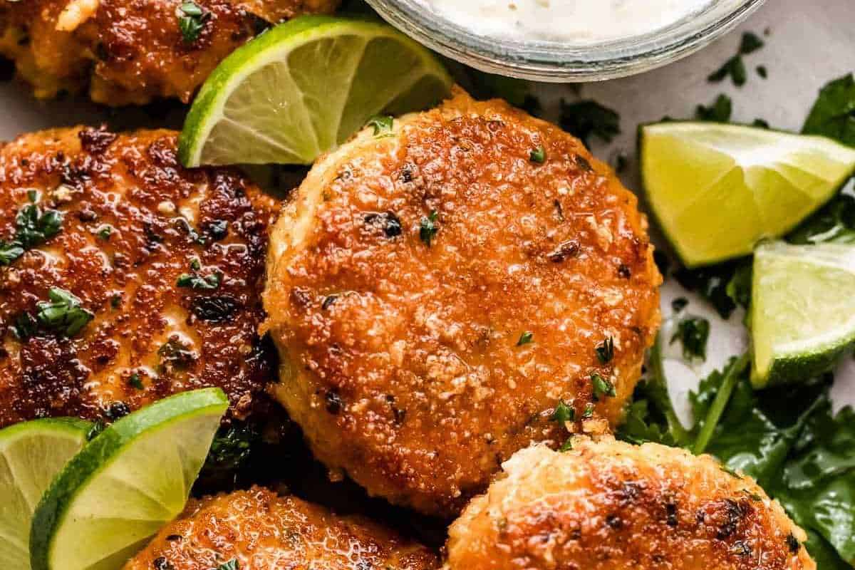 Golden-brown salmon patties served with lime wedges and a side of creamy sauce, garnished with chopped herbs.