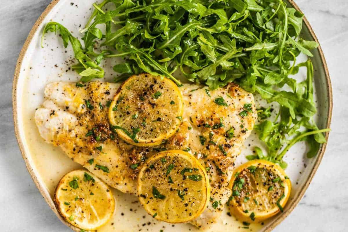 A plate of lemon chicken garnished with fresh parsley and thin lemon slices, accompanied by a side of arugula salad.