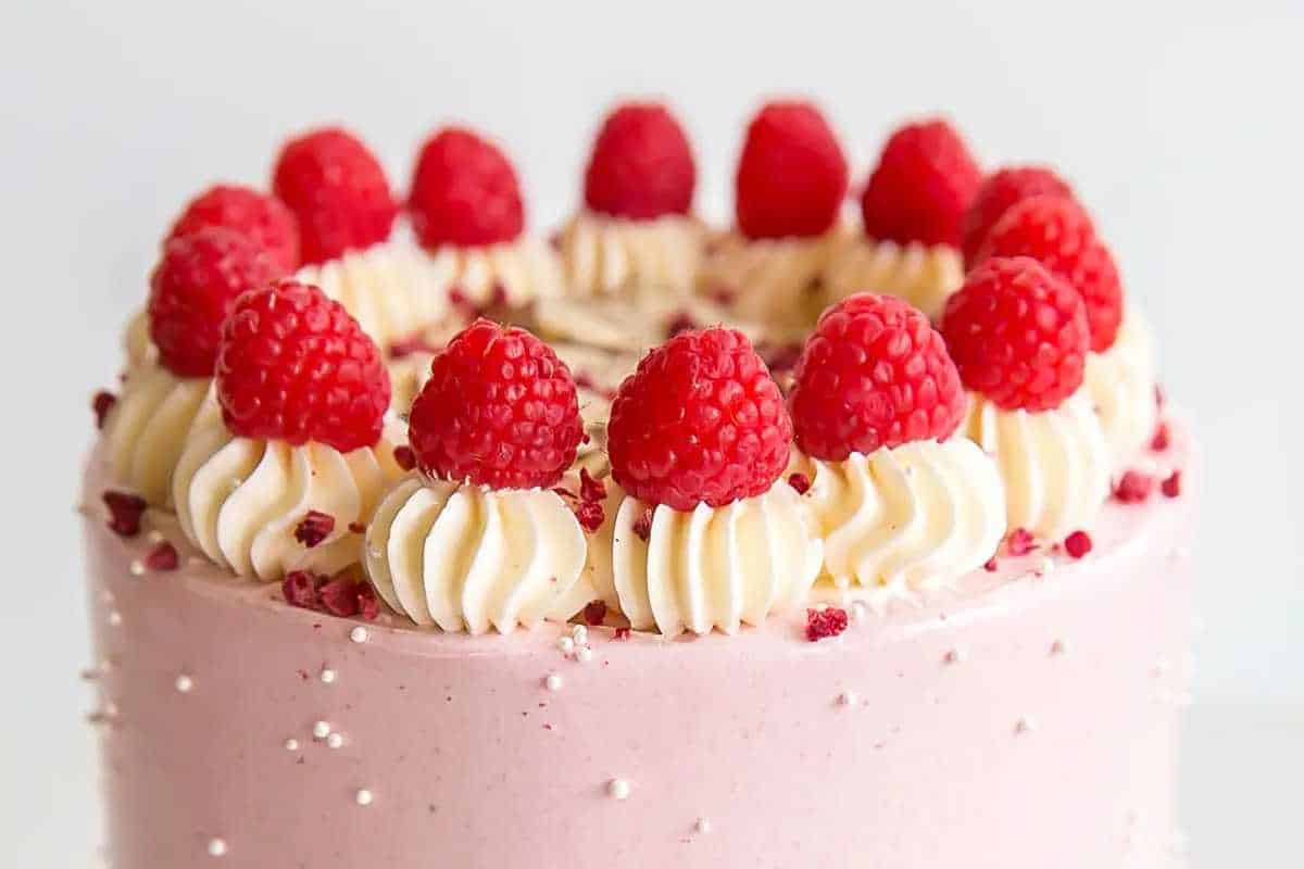 A pink frosted cake decorated with swirls of cream and topped with fresh raspberries.