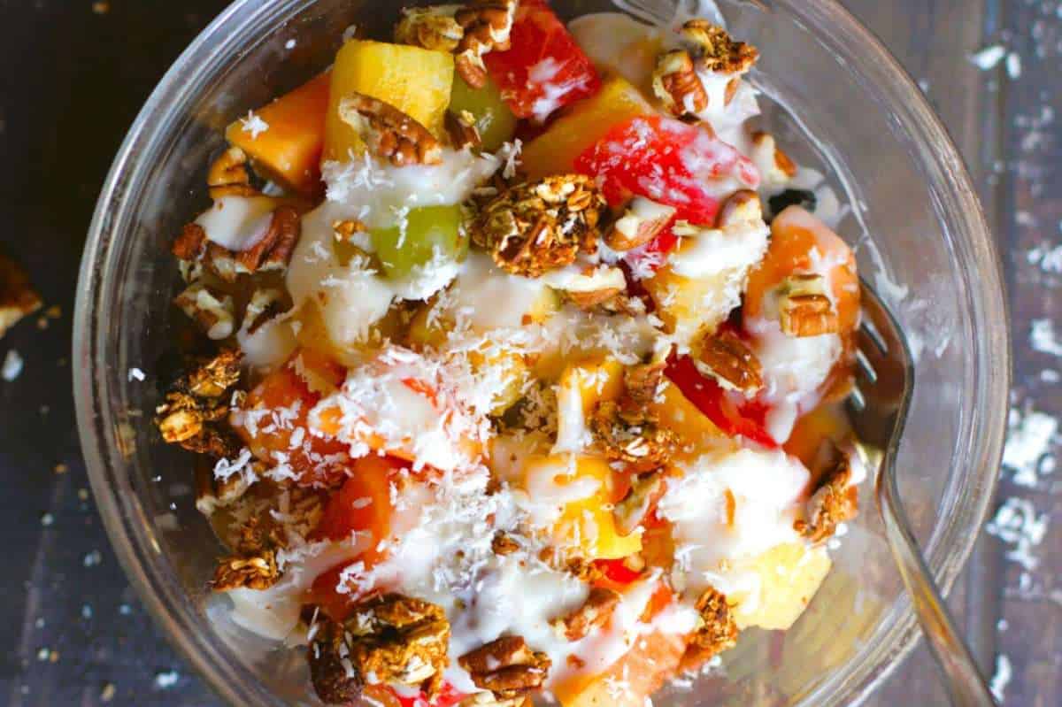 A bowl of tropical fruit salad topped with granola, coconut shavings, and a drizzle of honey, viewed from above.