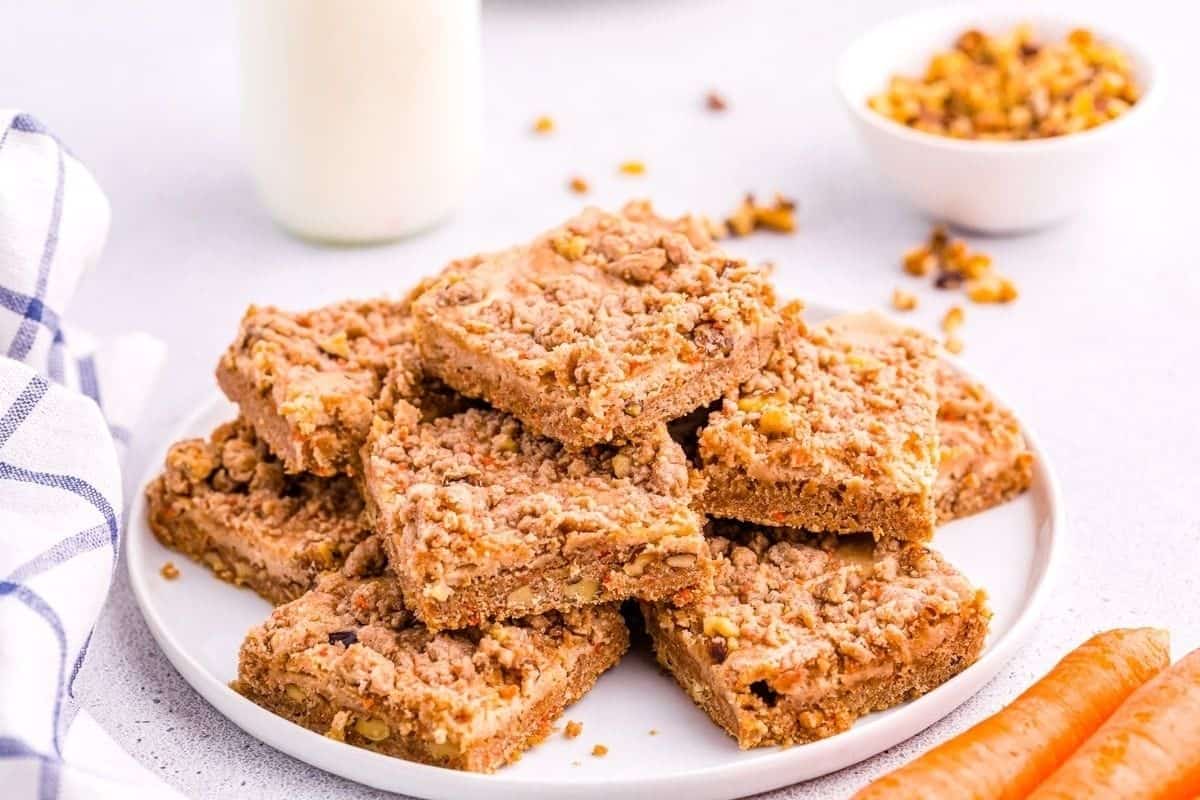 A plate of carrot cake bars with crumble topping accompanied by carrots and a small bowl of nuts.