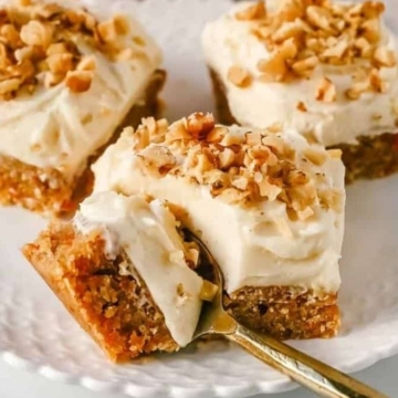 Three slices of carrot cake with cream cheese frosting and chopped nuts on a white plate with a gold fork.