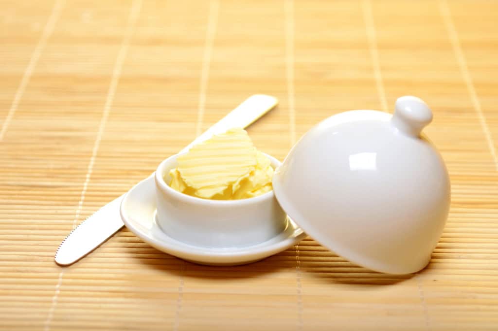 A small white dish containing butter with a silver knife next to it, and a ceramic lid resting on a bamboo mat.