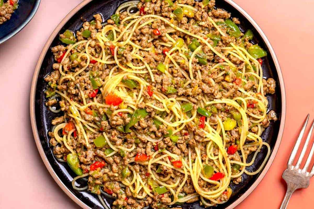 A skillet with spaghetti, ground meat, and mixed vegetables, served on a pink surface with a fork on the side.