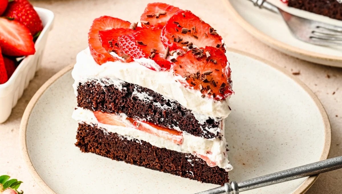 A slice of chocolate cake with cream and strawberries on a plate, surrounded by fresh strawberries.