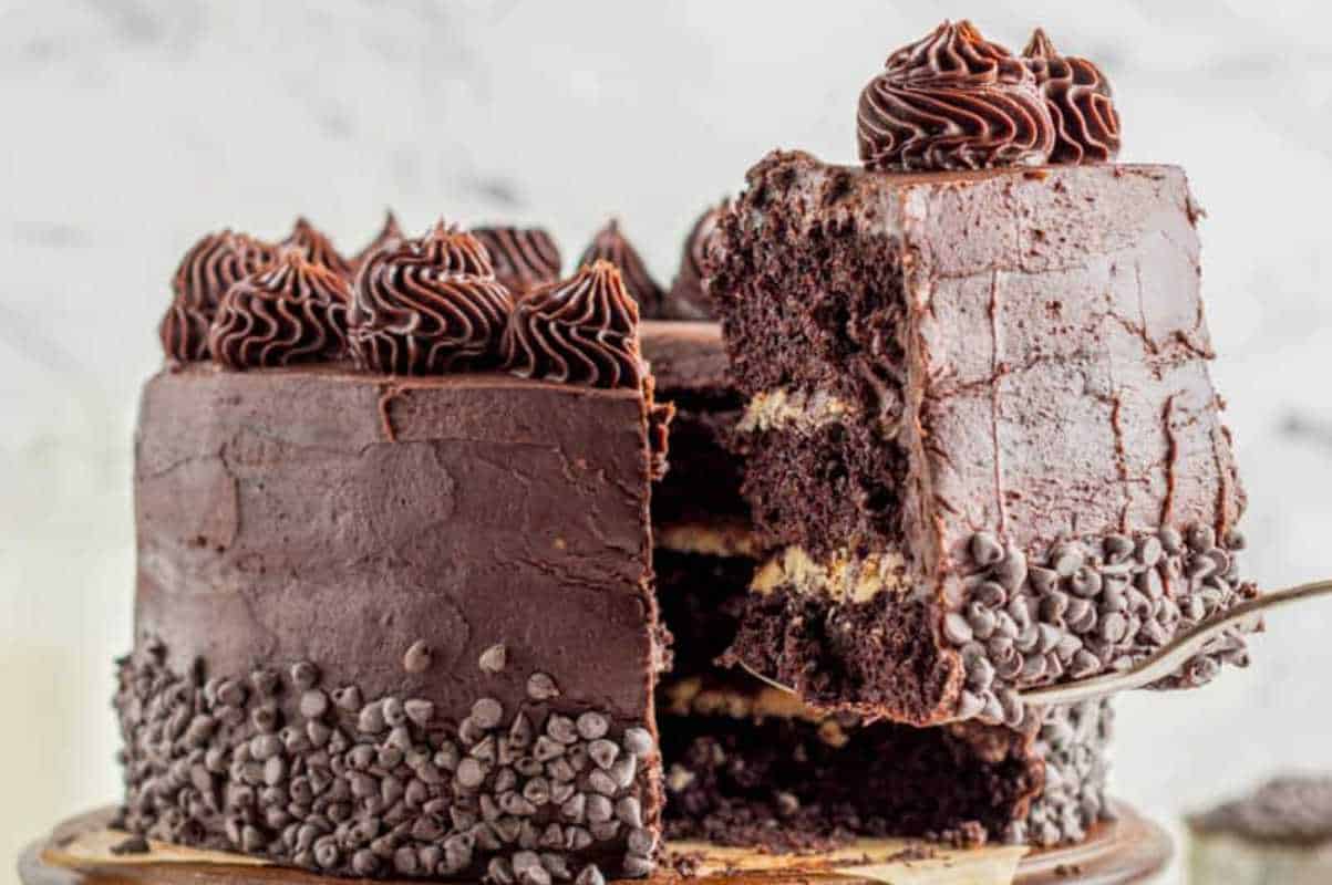 A layered chocolate cake with chocolate frosting and mini chocolate chips, a slice is being served with a cake server.