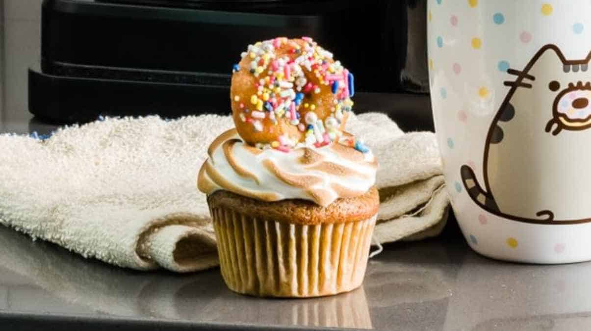 A frosted cupcake with sprinkles and a mini doughnut on top, sitting on a kitchen counter next to a mug with a cat illustration.