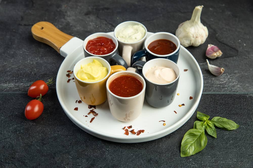 A variety of sauces in small cups on a white plate, accompanied by cherry tomatoes, garlic, and basil leaves on a dark surface.