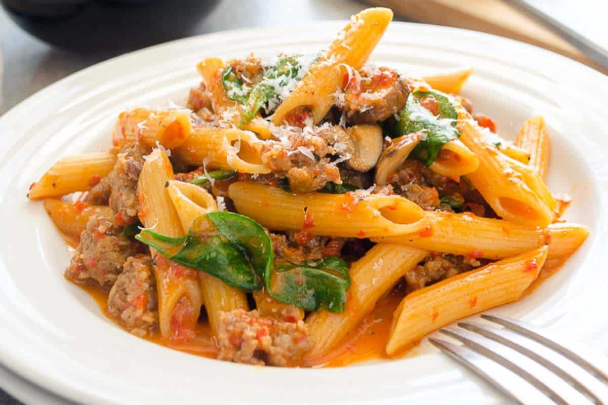 A plate of penne pasta with tomato sauce, ground meat, mushrooms, spinach, and grated cheese.