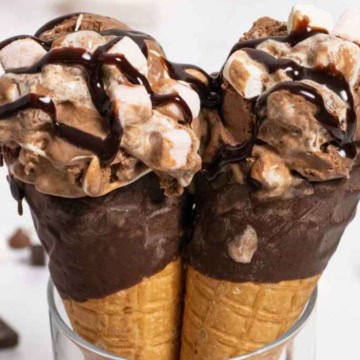 Two chocolate ice creams with marshmallows and chocolate sauce in waffle cones, placed in a glass.