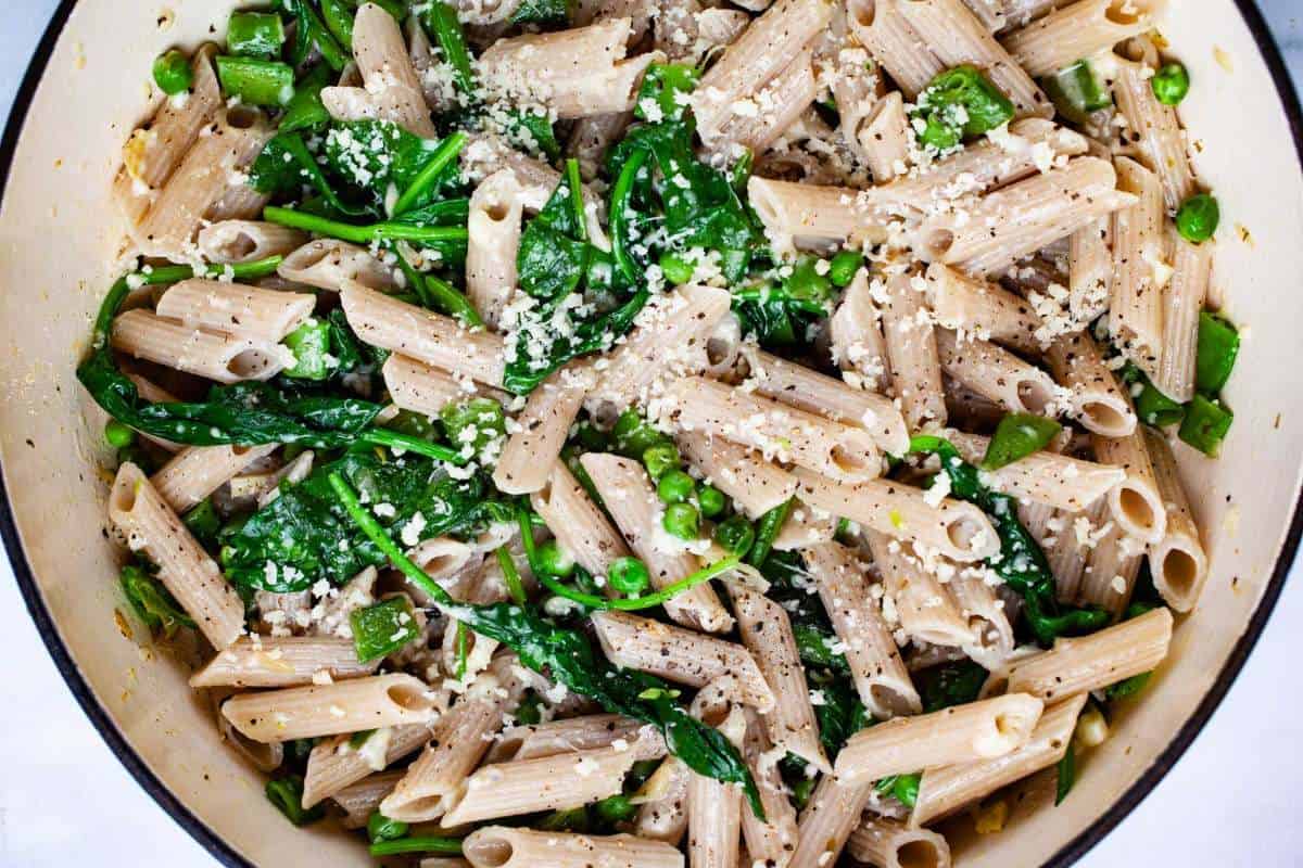 A pot of whole wheat penne pasta mixed with spinach, peas, and garnished with grated cheese.