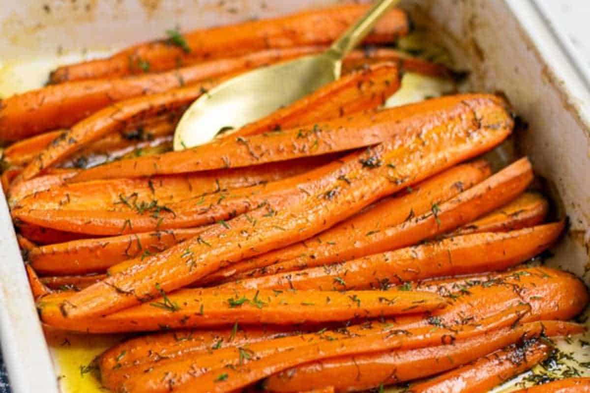 Roasted carrots with herbs in a baking dish, garnished with fresh dill and served with a golden serving spoon.