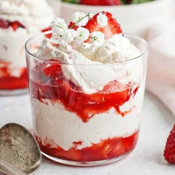 A parfait with layers of whipped cream and strawberry compote in a glass, garnished with fresh strawberries and small white flowers.