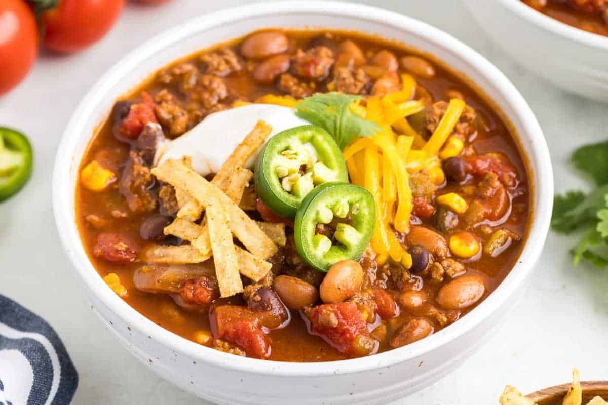 A bowl of chili topped with sour cream, shredded cheese, jalapeño slices, and tortilla strips, accompanied by fresh tomatoes.