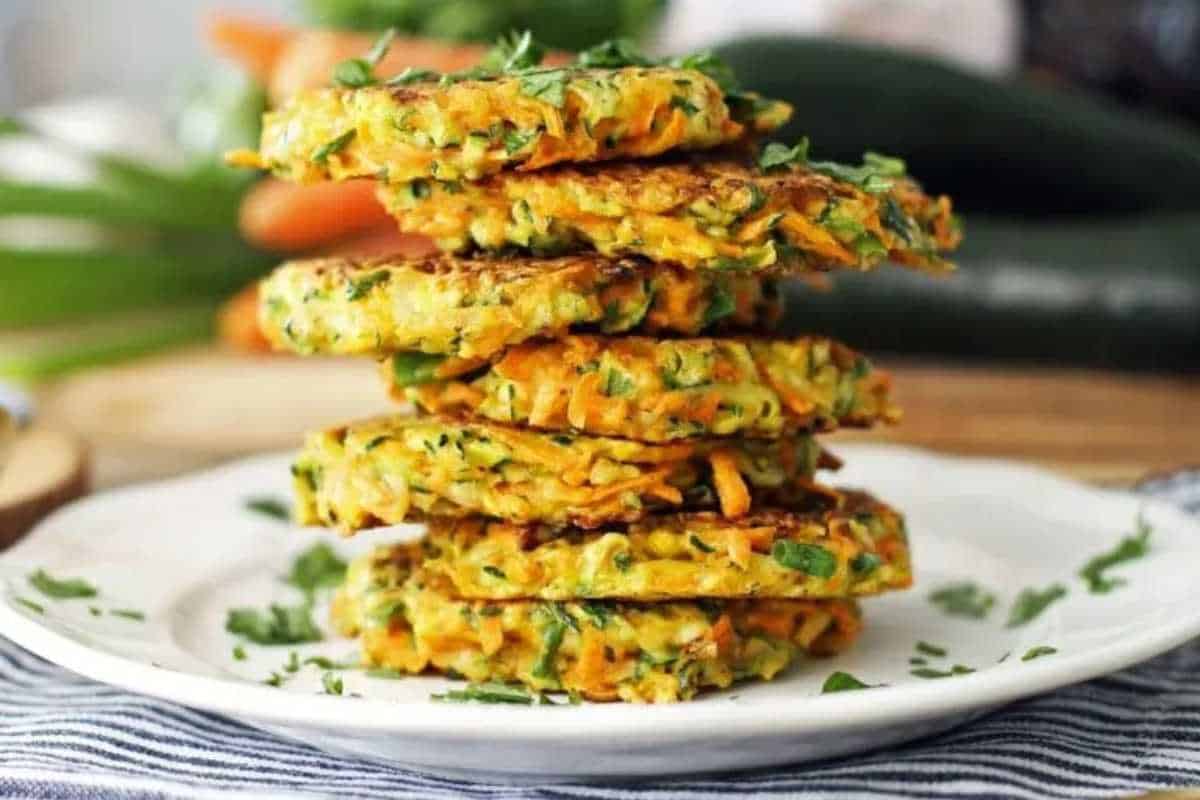 A stack of vegetable fritters on a white plate, garnished with herbs, with fresh zucchinis and carrots in the background.