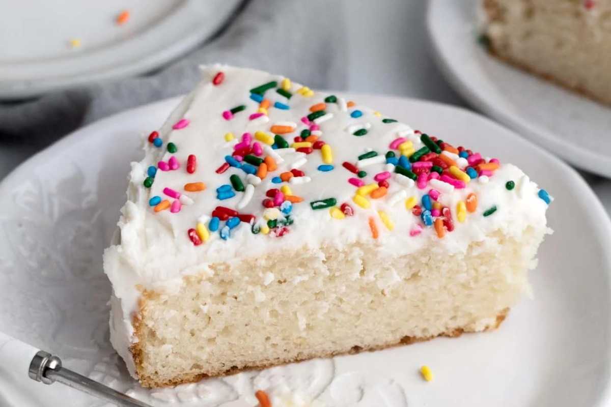 A slice of vanilla cake with white frosting and colorful sprinkles, served on a white plate with a fork on the side.