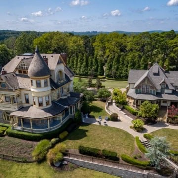 Aerial view of a luxurious estate featuring a large main house with a turret and a smaller house, surrounded by manicured gardens and a pool.