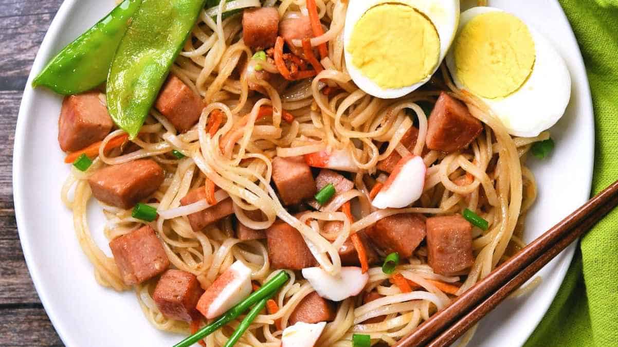 A plate of stir-fried noodles with diced sausage, sliced boiled eggs, and green peas, accompanied by chopsticks.