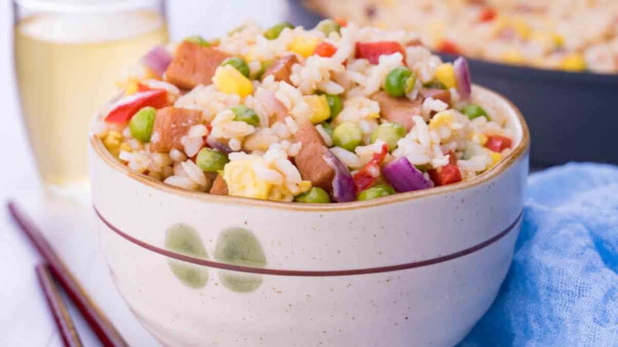 A bowl of fried rice with vegetables and ham, served with a drink and chopsticks on the side.