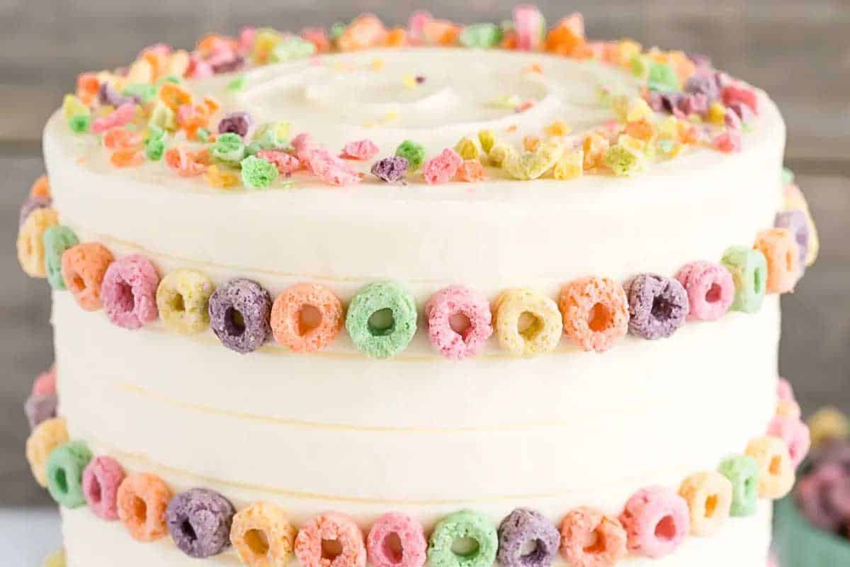 A two-tiered white frosted cake decorated with colorful cereal loops around each layer, displayed on a plain background.