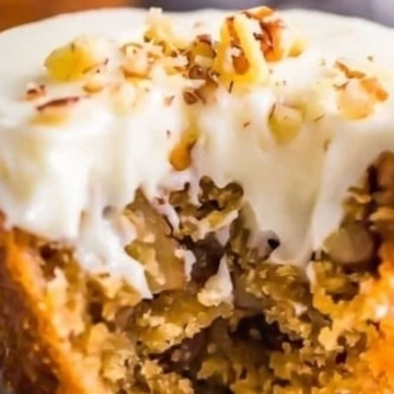 A carrot muffin with a bite taken out, topped with creamy white frosting and sprinkled with chopped nuts.