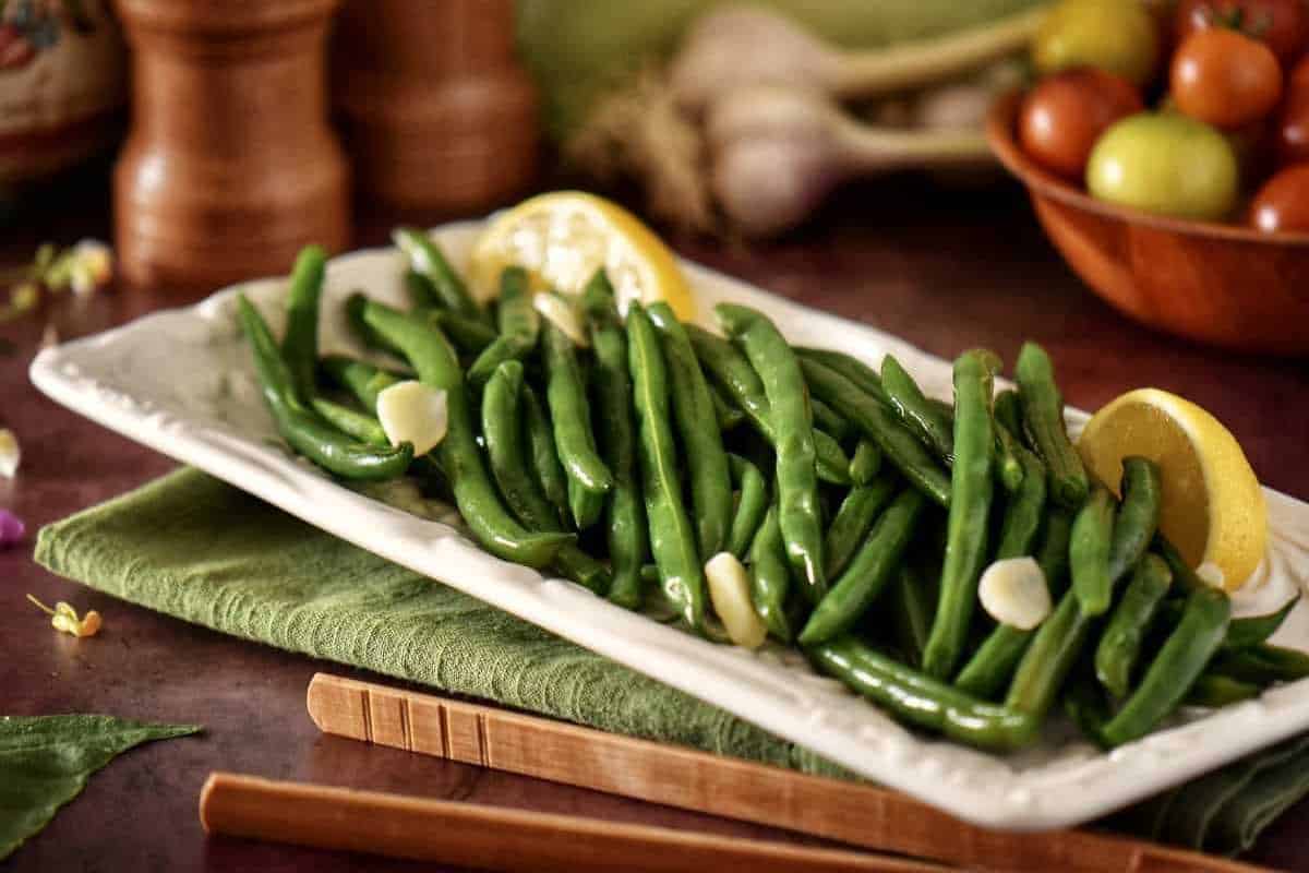 A plate of cooked green beans garnished with lemon slices and garlic, placed on a green napkin with various vegetables in the background.