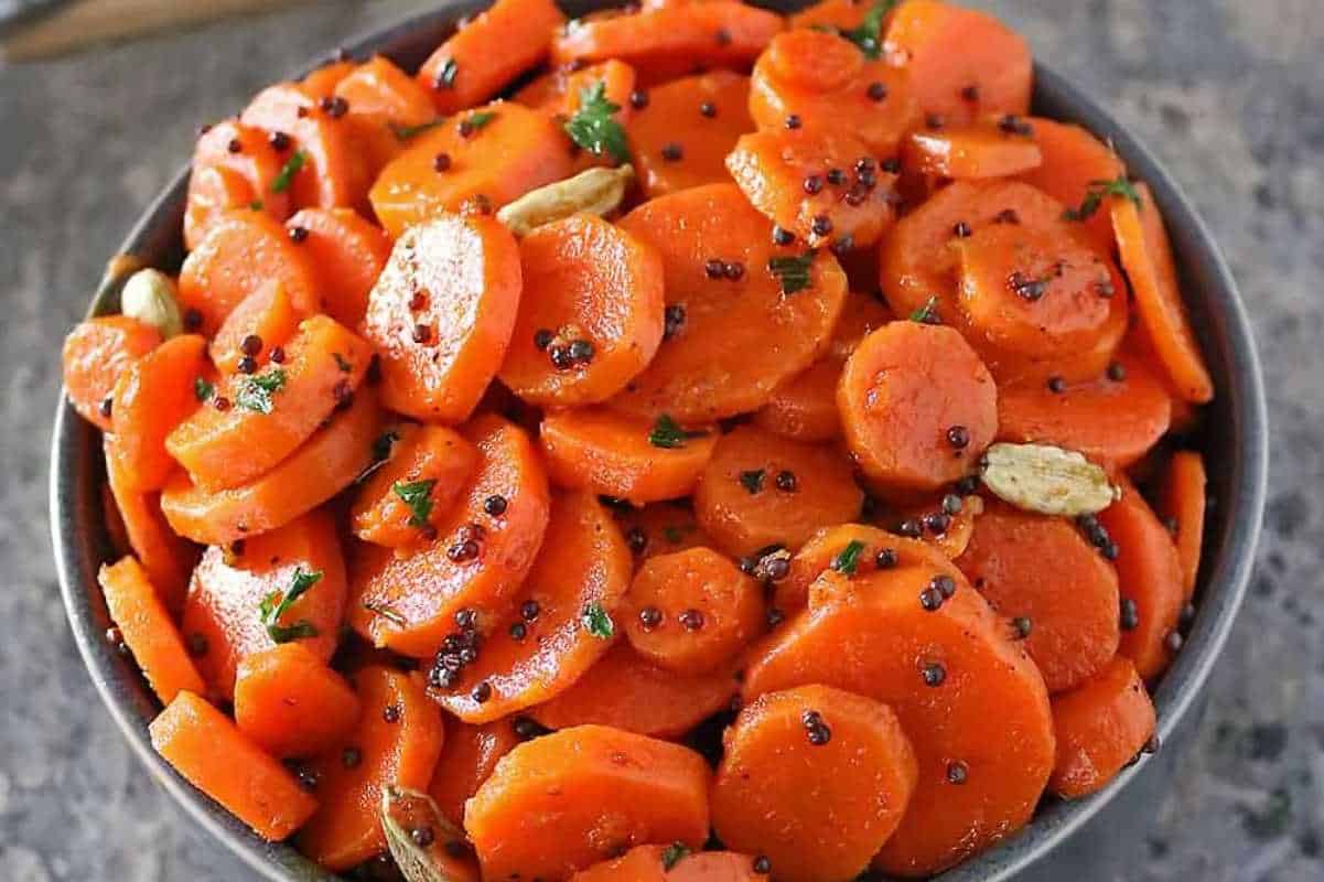 A bowl of sliced, glazed carrots garnished with parsley and black pepper, served with whole garlic cloves.