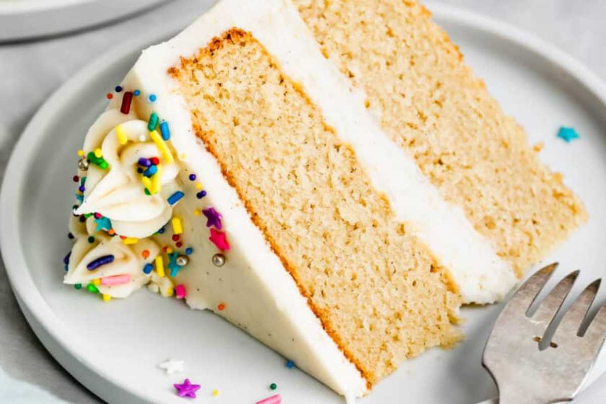 A slice of vanilla cake with white frosting and colorful sprinkles on a gray plate, accompanied by a fork on the side.