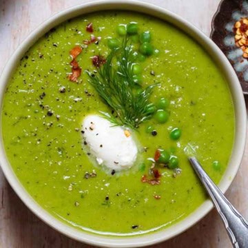 A bowl of vibrant green pea soup topped with a dollop of cream, fresh herbs, and bacon bits, served with a spoon on the side.