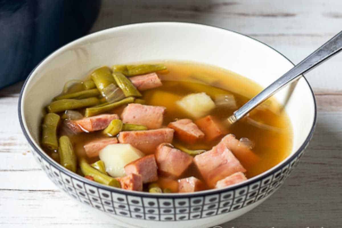 A bowl of soup with green beans, diced potatoes, and sliced sausage, served with a spoon on a wooden table.