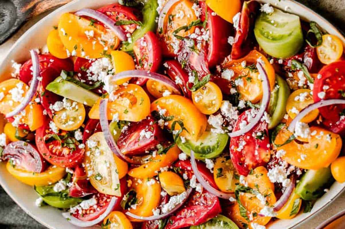 A colorful tomato salad with red onions, feta cheese, and herbs in a white bowl.