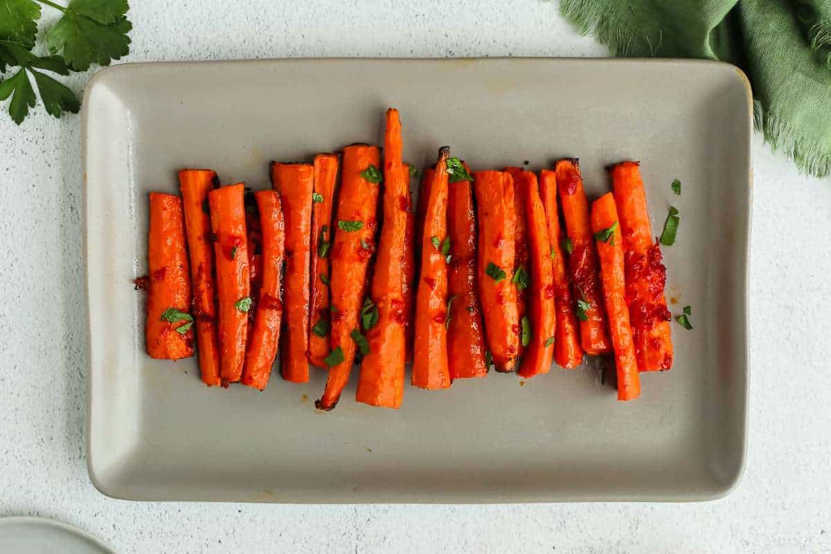 A rectangular plate containing seasoned roasted carrots garnished with herbs, served on a white tabletop with a green napkin on the side.