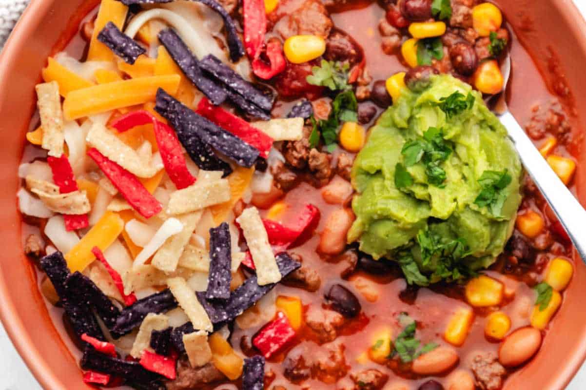 A bowl of chili with corn, beans, cheese, tortilla strips, and a dollop of guacamole.