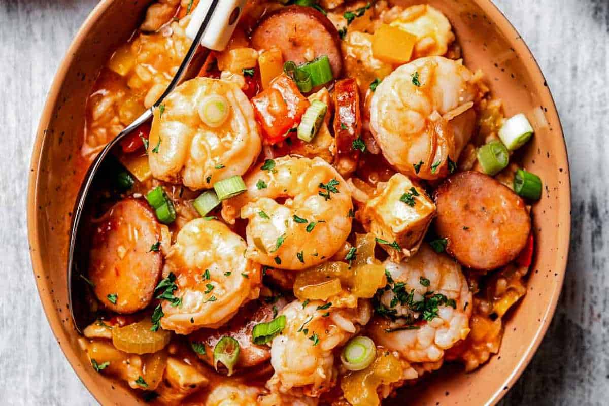 A bowl of shrimp and sausage gumbo with chunks of tofu, chopped vegetables, and a sprinkling of herbs, served with a spoon.