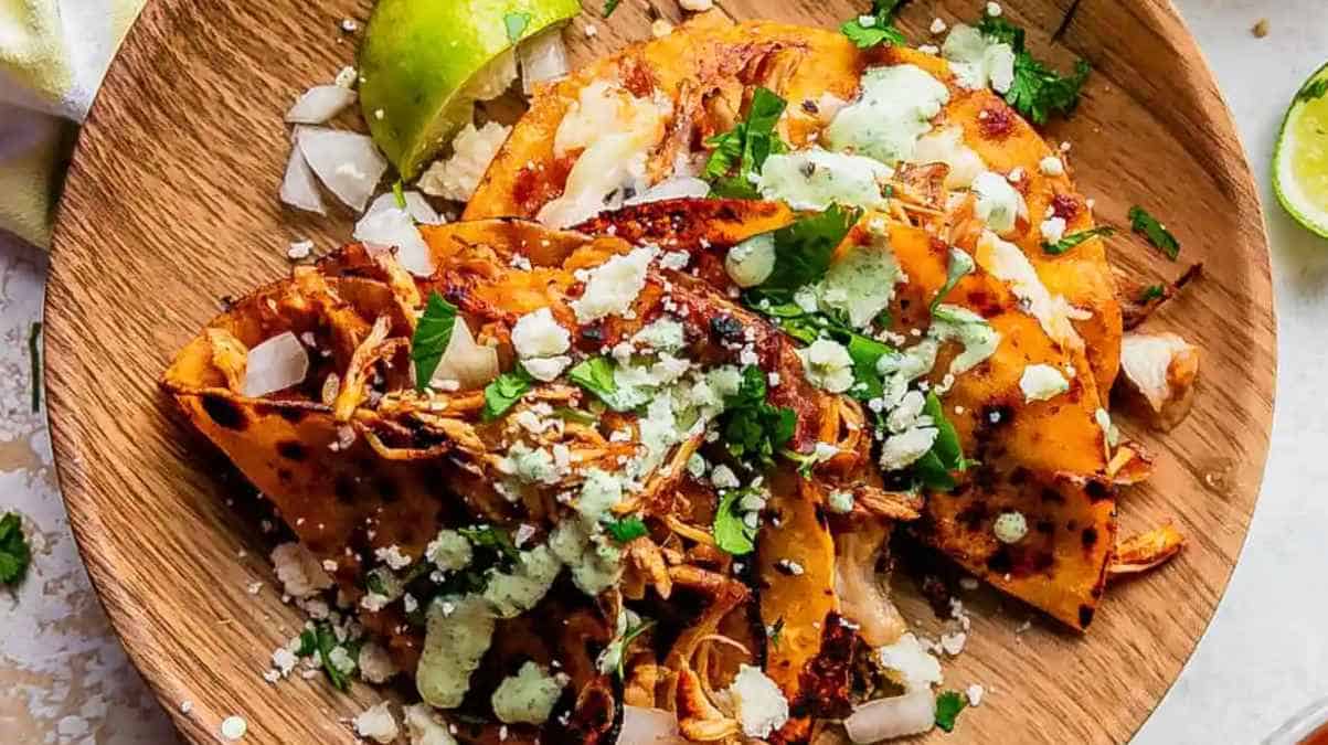 A wooden plate of grilled tacos topped with avocado slices, crumbled cheese, cilantro, and a lime wedge on the side.