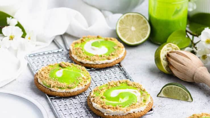 Lime-glazed cookies on a cooling rack with a jar of green icing and baking utensils on a kitchen counter.