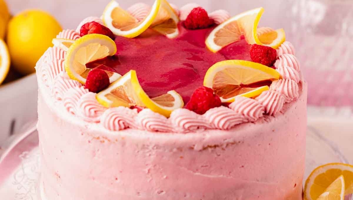 A pink frosted cake decorated with lemon slices and raspberries on a glass stand.