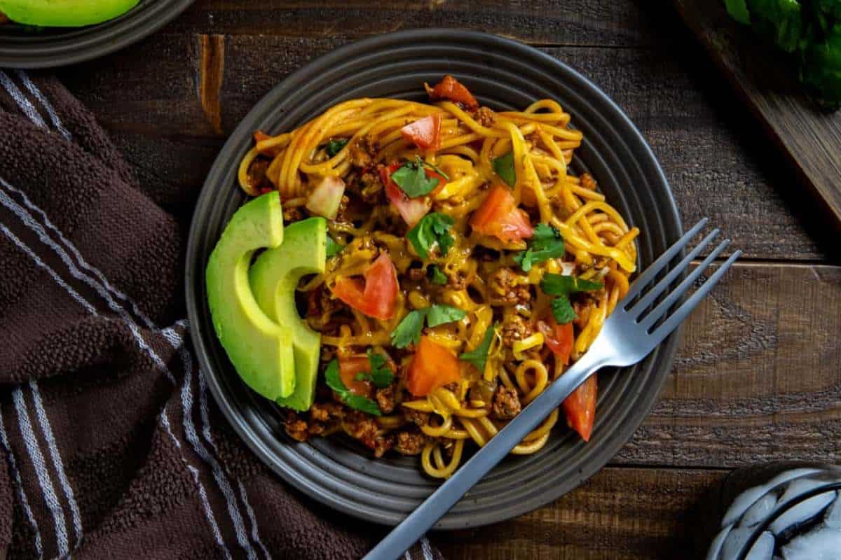 A plate of spaghetti with tomato, ground meat, and slices of avocado, accompanied by a fork.