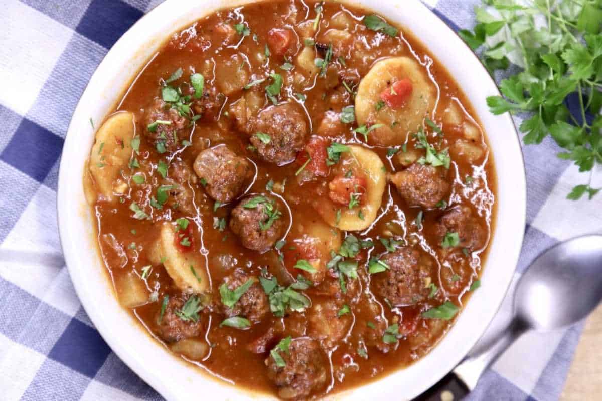 A bowl of meatball and potato stew garnished with chopped parsley, served on a blue and white checkered cloth.