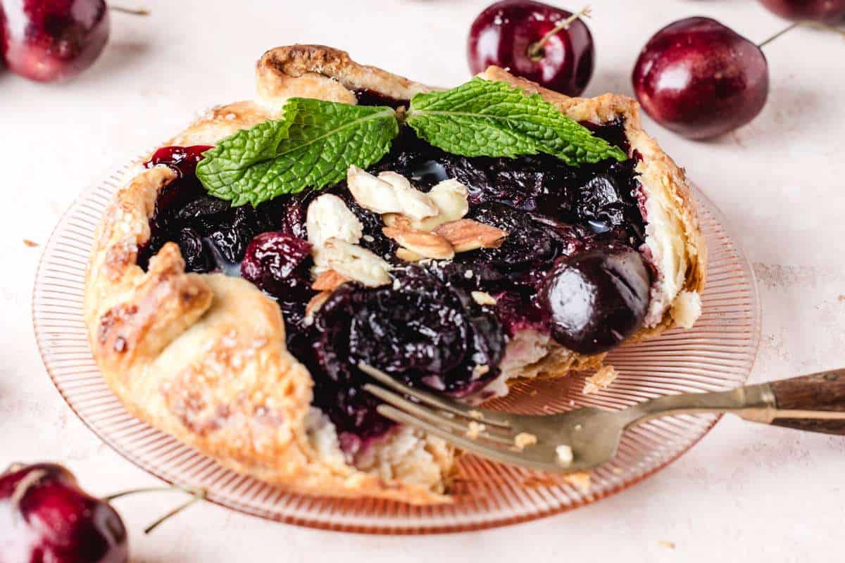 A slice of cherry pie topped with almonds and mint leaves on a plate, with fresh cherries around it and a fork on the side.