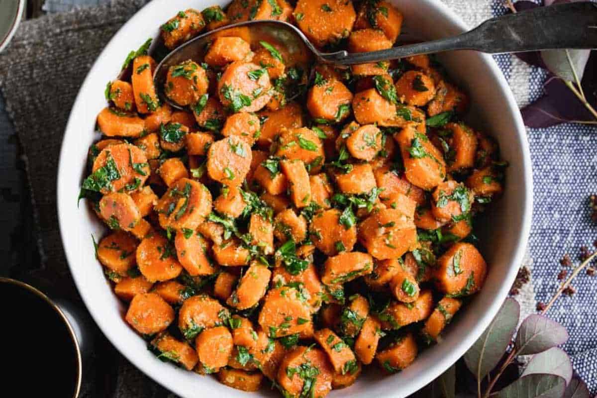 A bowl of sliced glazed carrots topped with chopped parsley, with a serving spoon inside, placed on a textured grey cloth.