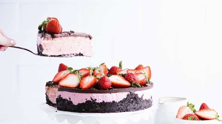 A strawberry cheesecake with a chocolate crust, garnished with fresh strawberries, with a slice being served.