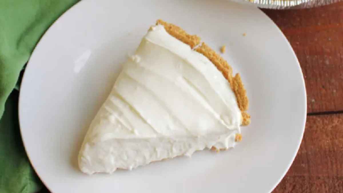 A slice of classic cheesecake with a smooth, white topping and a golden crust, served on a white plate with a green napkin beside it.