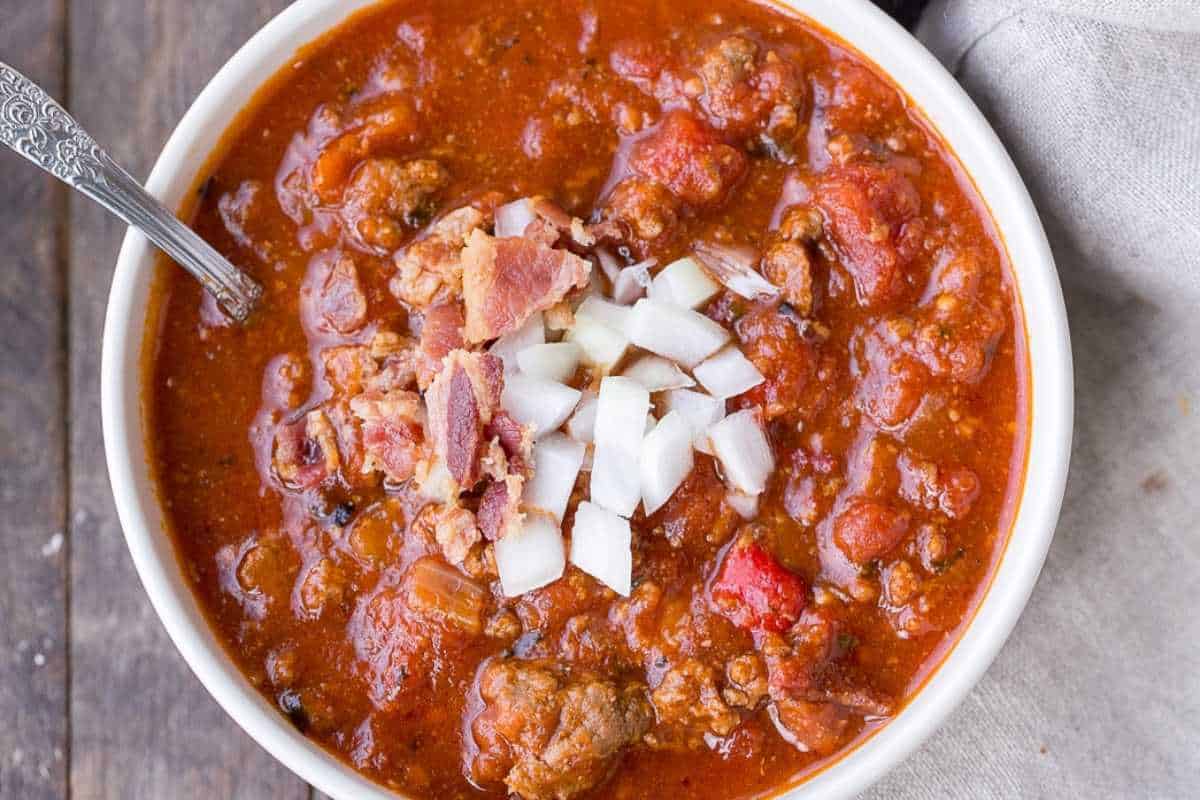 A bowl of chili with ground meat, tomatoes, and diced onions, topped with pieces of bacon, served on a wooden surface.