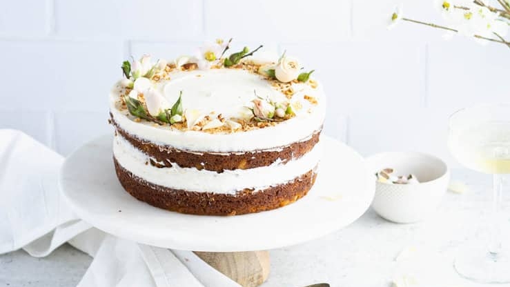 A two-layer naked cake with white frosting and decorated with nuts and small flowers, presented on a wooden stand with plates and cutlery on the side.