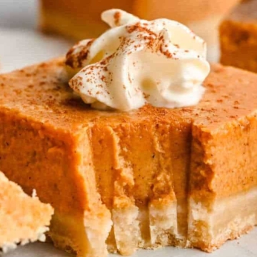 A slice of pumpkin cheesecake topped with whipped cream and a sprinkle of cinnamon, with more slices in the background.