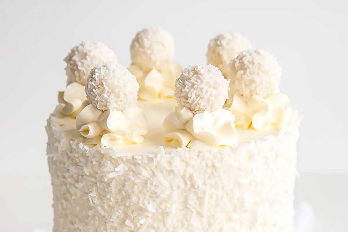 White coconut cake decorated with cream swirls and coconut truffles on top, set against a plain white background.