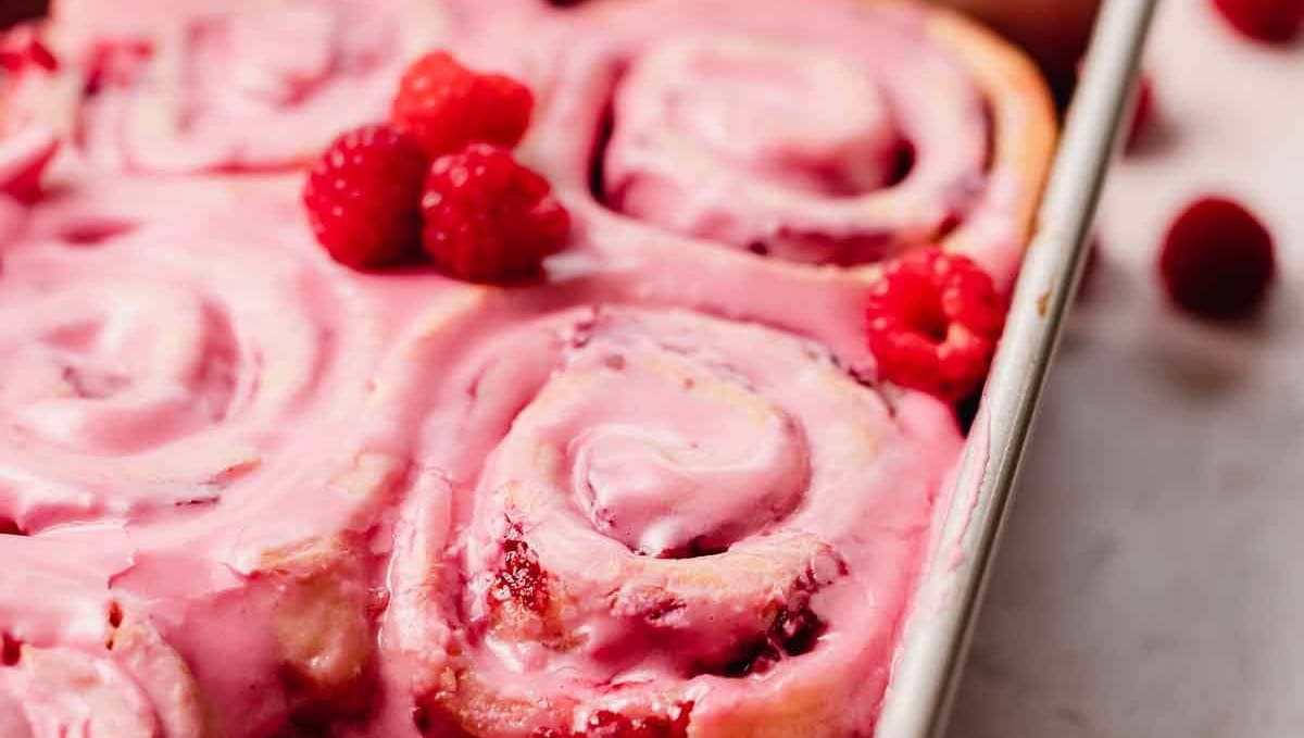 A pan of freshly baked raspberry rolls with pink icing and whole raspberries on top.