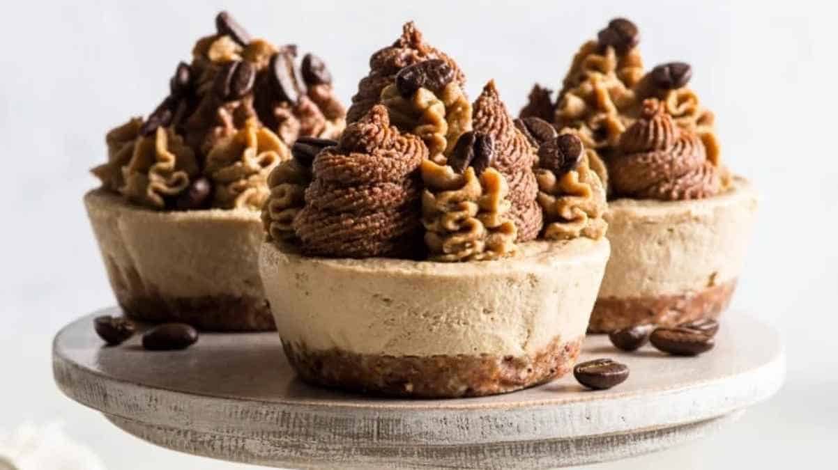 Three coffee-flavored mini cheesecakes garnished with coffee beans and piped cream on a white stand.